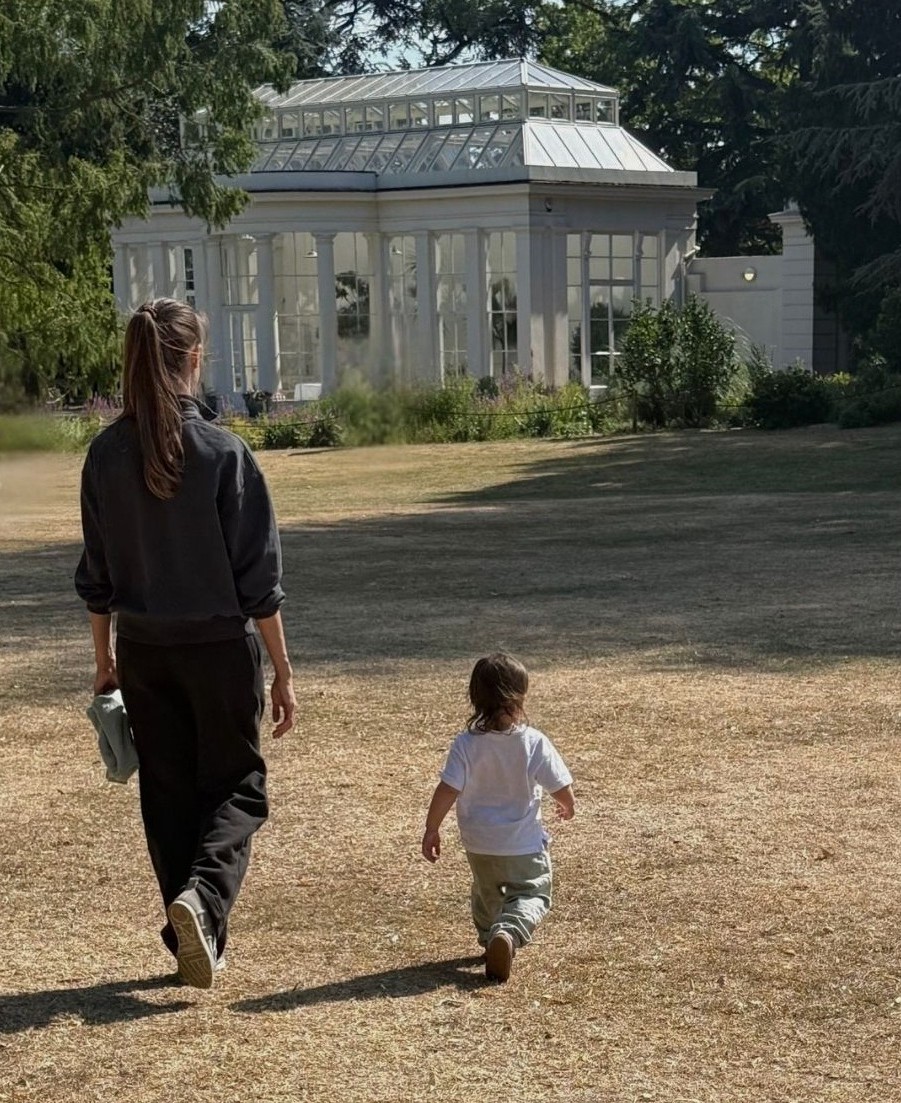 Anastasia walking with Zoe across a sunlit park lawn towards a white glasshouse